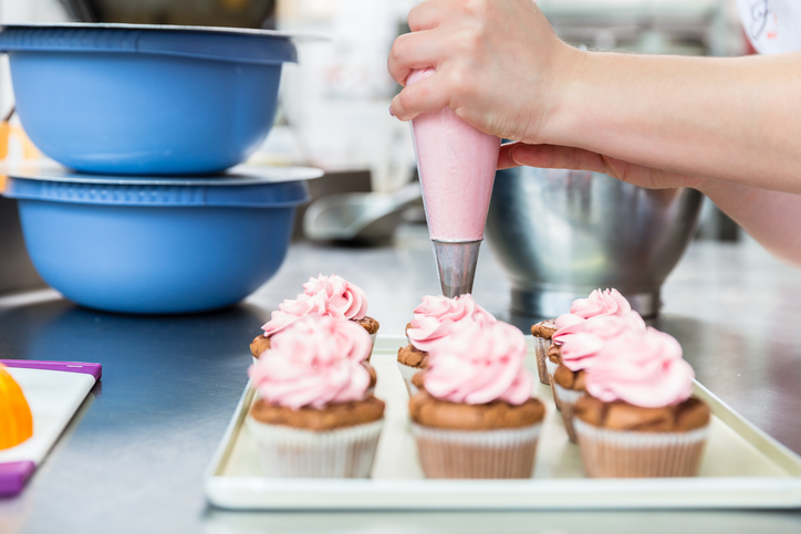 Mit einem Spritzbeutel wird eine gleichmäßige Haube aus rosa Himbeercreme auf die Schokocupcakes gespritzt.