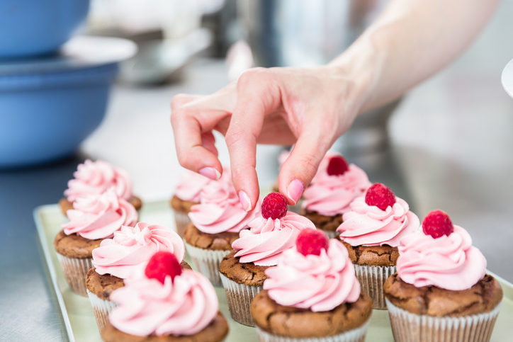 Eine Hand setzt vorsichtig eine frische Himbeere auf das rosa Frosting eines Schokocupcakes.