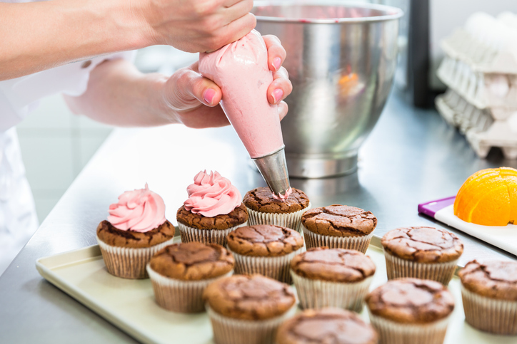 Nahaufnahme beim Auftragen der rosa Creme auf frisch gebackene dunkle Schokoladenmuffins.