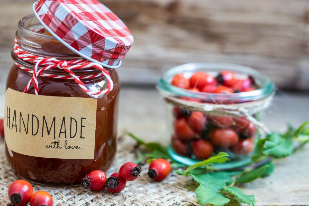 Ein Glas mit Hagebuttenmarmelade und der Aufschrift „HANDMADE with love“ steht neben einem kleinen Glas mit frischen Hagebutten auf einem Holztisch.