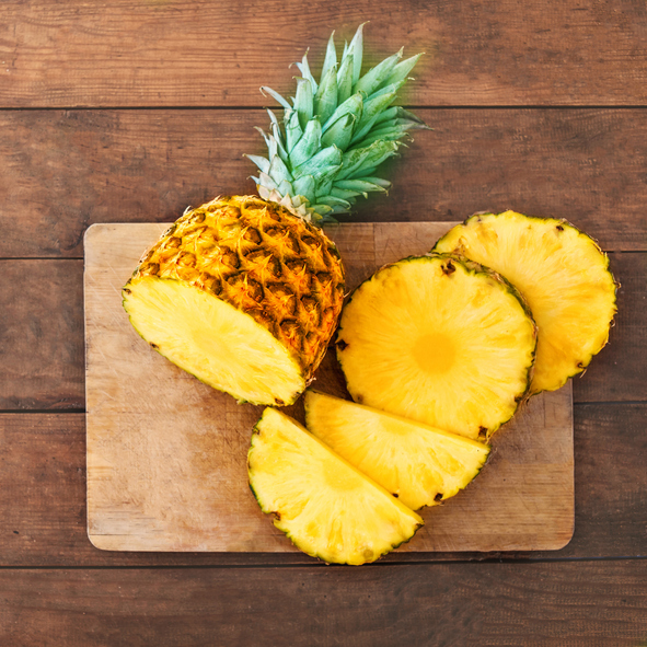 Von oben fotografierte Ananas-Hälfte und perfekt geschnittene Ringe auf einem hellen Holzschneidebrett.