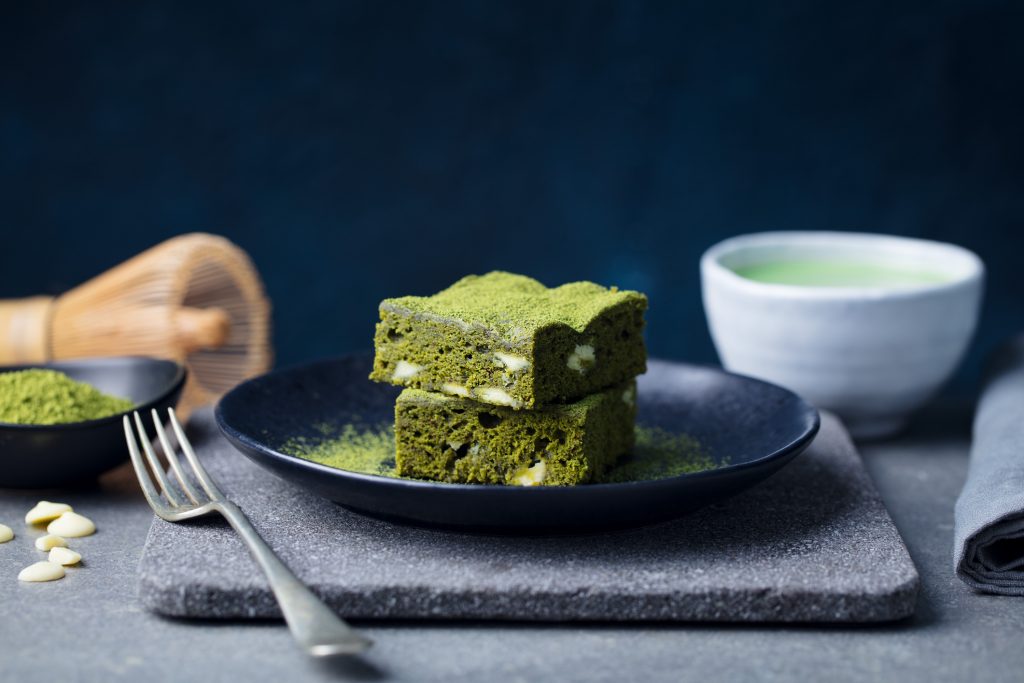 Zwei übereinander gestapelte, leuchtend grüne Matcha-Brownie-Stücke liegen auf einem dunklen Teller, bestäubt mit feinem Matcha-Pulver, begleitet von einem Matchatee und Backutensilien im Hintergrund.