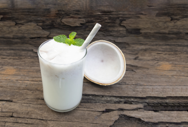 Ein Glas mit einem weißen Kokos-Shake, garniert mit frischer Minze und einem Strohhalm, steht neben einer halbierten Kokosnuss auf einem rustikalen Holzuntergrund.
