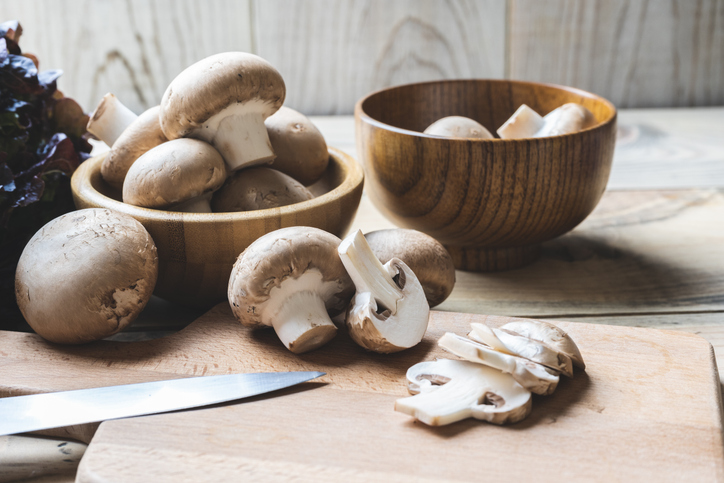 Eine Auswahl an ganzen und bereits in Scheiben geschnittenen braunen Champignons liegt in und neben Holzschalen auf einem Schneidebrett.