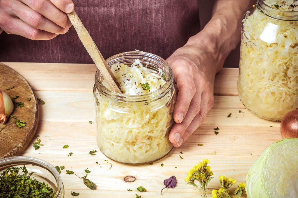 Hände drücken Sauerkraut mit einem Holzlöffel in ein Glas, neben dem ein weiteres volles Glas und frische Zutaten auf einem Holztisch stehen.