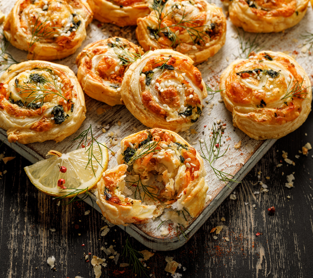 Goldbraun gebackene, schneckenförmige Blätterteig-Pinwheels mit Lachs, verziert mit frischem Dill und Sesamkörnern, serviert auf einem rustikalen Holzbrett mit einer Zitronenscheibe