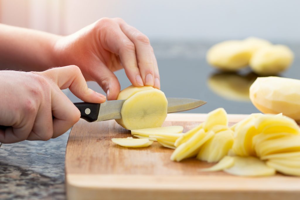 Hände schneiden geschälte Kartoffeln mit einem Messer auf einem Holzbrett in feine Scheiben.