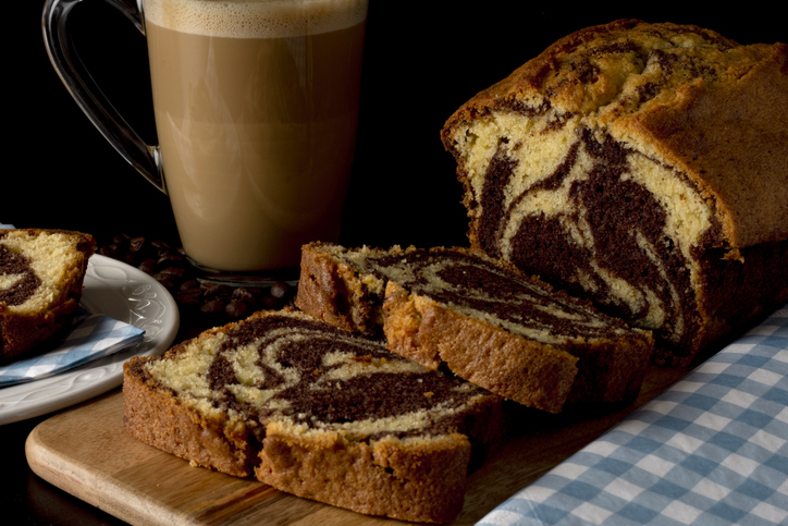 Ein aufgeschnittener Marmorkuchen liegt auf einem Holzbrett neben einer Tasse Kaffee.