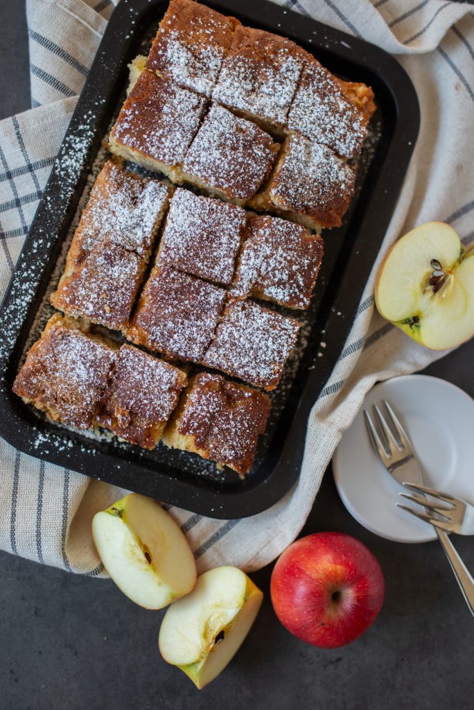 Ein auf einem Backblech in gleichmäßige Stücke geschnittener Apfelkuchen, der großzügig mit Puderzucker bestäubt ist, flankiert von frischen Apfelstücken.