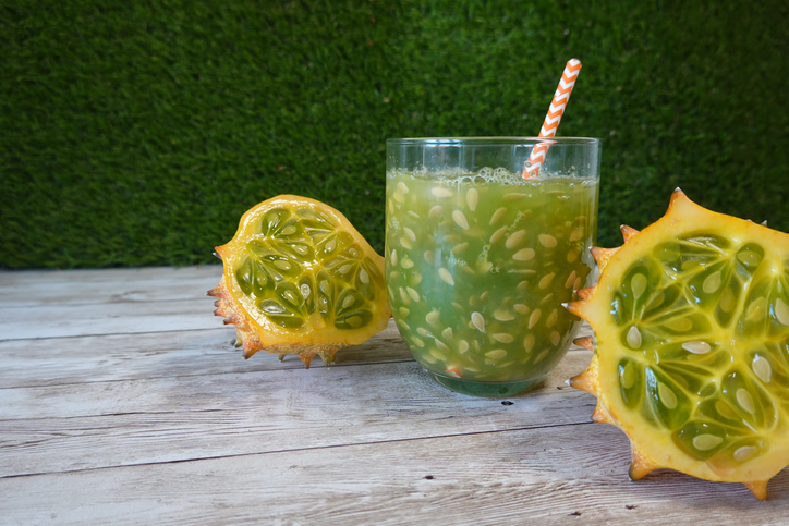 Ein Glas mit Kiwano-Saft flankiert von zwei aufgeschnittenen Kiwano-Früchten auf einer Holzoberfläche.