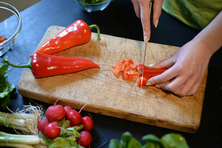 Hände schneiden eine rote, längliche Paprika auf einem hölzernen Schneidebrett in kleine Stücke.