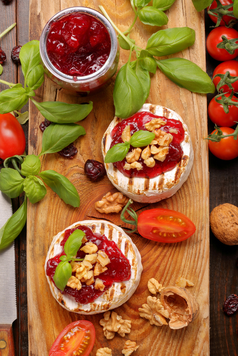 Zwei gegrillte Camembert-Käse, garniert mit Preiselbeeren, Walnüssen und Basilikum auf einem rustikalen Holzbrett.