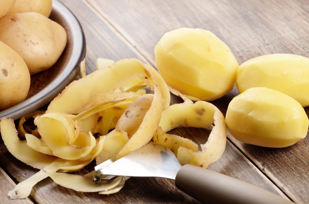 Geschälte Kartoffeln, Kartoffelschalen und ein Sparschäler liegen auf einem rustikalen Holztisch.