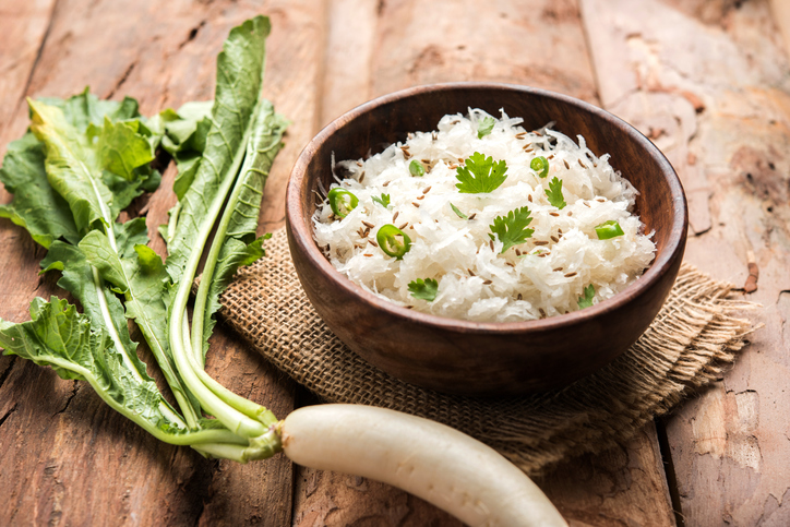 Ein frisch zubereiteter, fein geraspelter Rettichsalat mit Kräutern und Gewürzen in einer rustikalen Holzschale, daneben liegt ein ganzer Rettich mit grünen Blättern.