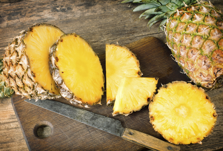 Eine aufgeschnittene Ananas und frische Scheiben liegen auf einem dunklen Holzbrett neben einem alten Messer.