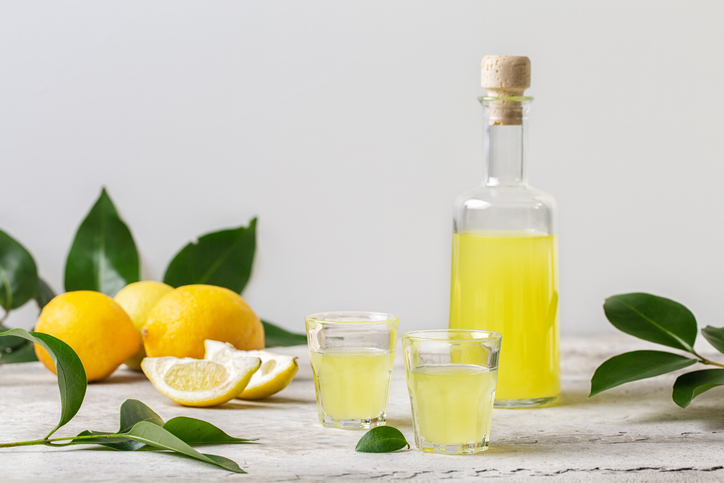 Eine Flasche Limoncello, kleine Gläser und frische Zitronen stehen auf einem hellen Tisch.