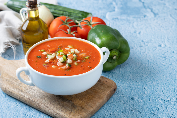 Eine Schale Gazpacho mit Tomaten, Gurke, Paprika und Olivenöl steht auf einem Holzbrett.