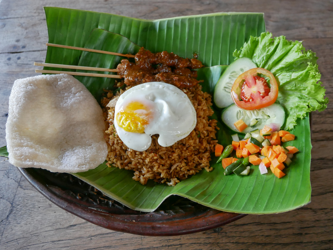 Nasi Goreng mit Spiegelei, Satay-Spießen, Krupuk und frischem Gemüse liegt auf einem Bananenblatt.