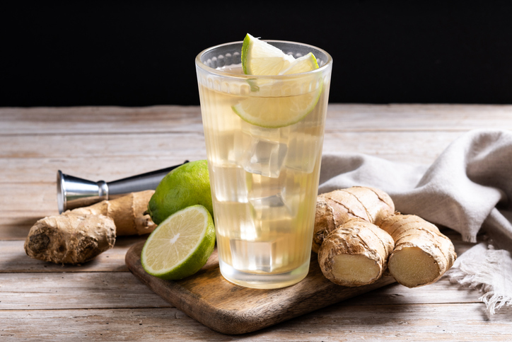 Ein Glas Ginger Ale mit Eiswürfeln, Limette und frischem Ingwer steht auf einem Holztisch.