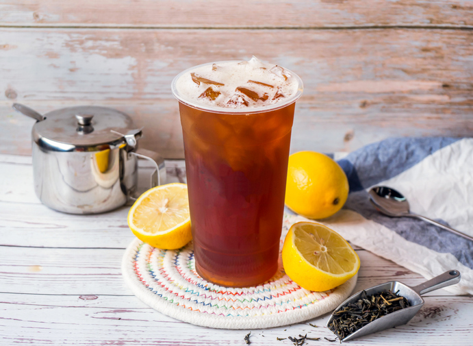 Glas mit schwarzem Eistee, Eiswürfeln und Zitronen auf einem hellen Holztisch.