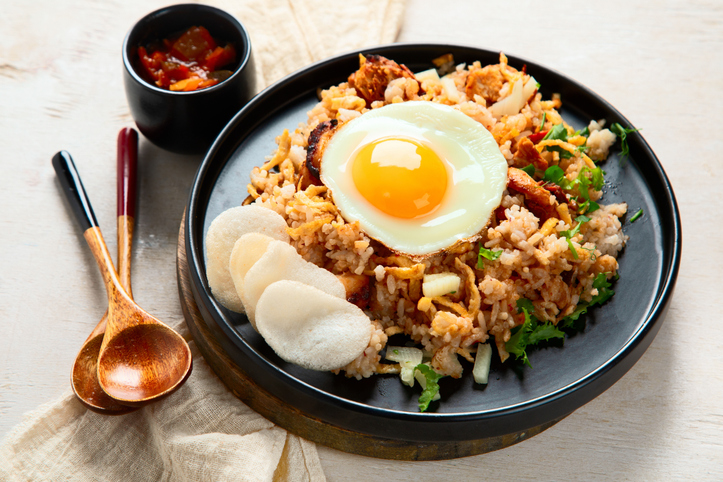 Nasi Goreng mit Spiegelei, Krupuk, Kräutern und würziger Sauce wird auf einem schwarzen Teller serviert.