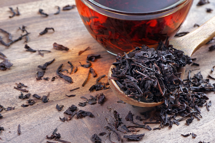 Lose schwarze Teeblätter auf einem Holzlöffel neben einer Tasse mit aufgebrühtem Tee.