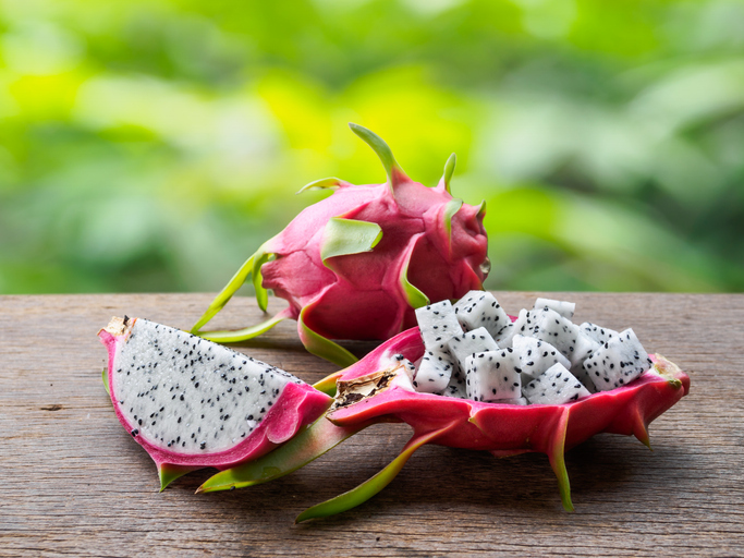 Eine aufgeschnittene Pitahaya mit weißen Fruchtwürfeln und schwarzen Kernen liegt auf einem Holztisch.