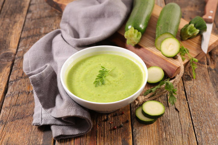 Hellgrüne Zucchinisuppe in einer weißen Schale mit Petersilie, frischen Zucchini und geschnittenen Zucchinischeiben auf einem Holztisch.