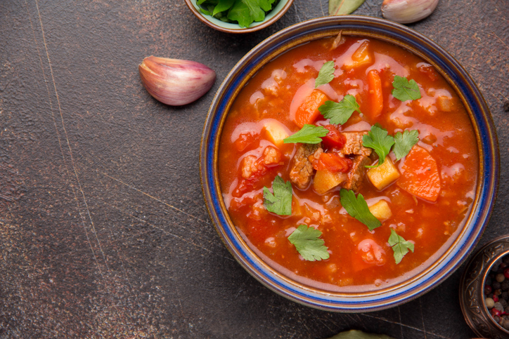 Gulaschsuppe von oben in einer Schale mit Fleisch, Kartoffeln, Karotten, Tomaten und frischen Kräuterblättern.