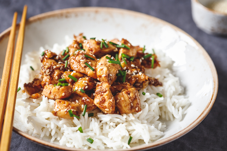 Teriyaki-Hühnchen mit Reis in einer Schale, garniert mit Sesam und fein geschnittenem Schnittlauch, daneben liegen Essstäbchen.