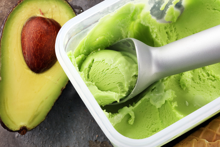 Cremiges Avocado-Eis in einer rechteckigen Eisbox mit Eislöffel, daneben liegt eine halbierte Avocado.