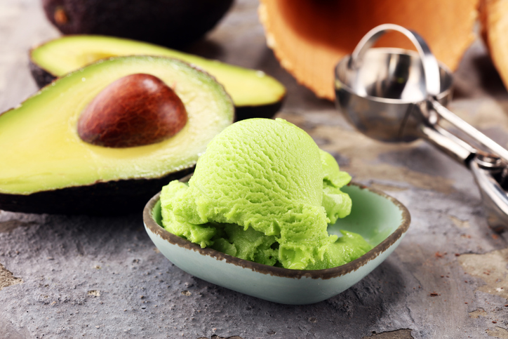 Eine Kugel Avocado-Eis in einer kleinen Schale, daneben liegen eine halbierte Avocado und ein Eisportionierer.