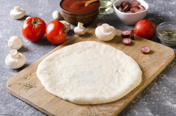 Runder Pizzateig auf einem Holzbrett mit Tomaten, Champignons, Wurststücken, Tomatensoße und Gewürzen im Hintergrund.