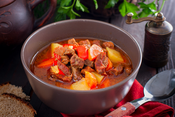 Gulaschsuppe in einer grauen Schüssel mit Rindfleisch, Kartoffeln, Paprika und Möhren auf einem dunklen Tisch.