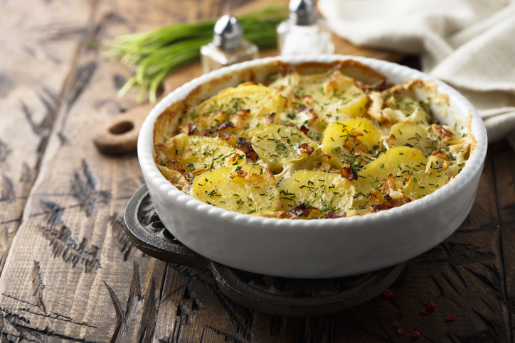 Kartoffel-Gratin in einer runden weißen Auflaufform mit goldgelben Kartoffelscheiben, cremiger Sauce und frischen Kräutern auf einem Holztisch.