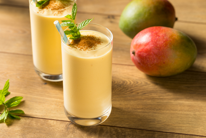 Zwei Gläser Mango Lassi mit Minze und Trinkhalmen auf einem Holztisch, daneben liegen zwei reife Mangos.