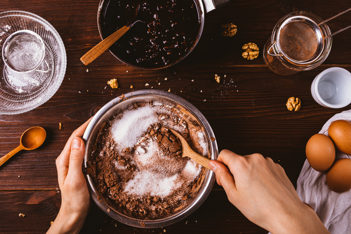 Schokoladiger Teig für Schoko-Cookies wird in einer Schüssel mit Zucker verrührt, daneben liegen Eier, Walnüsse und Küchenutensilien.