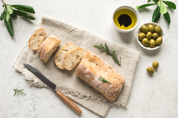 Aufgeschnittenes Ciabatta mit luftiger Krume neben Oliven, Olivenöl, Rosmarin und einem Brotmesser auf einer hellen Unterlage.