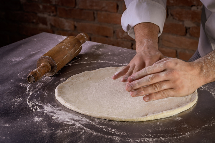 Hände formen einen runden Pizzateig auf einer bemehlten Arbeitsfläche, daneben liegt ein hölzernes Nudelholz.