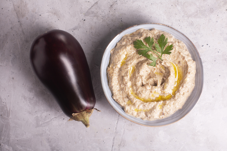 Heller Auberginen-Dip in einer Schale mit etwas Olivenöl und Petersilie, daneben liegt eine ganze Aubergine auf hellem Hintergrund.