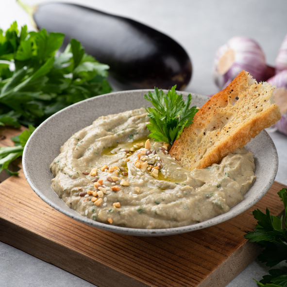 Cremiger Auberginen-Dip in einer grauen Schale mit Brot, Petersilie, Sesam und gehackten Nüssen, im Hintergrund liegen Aubergine und Zwiebeln.