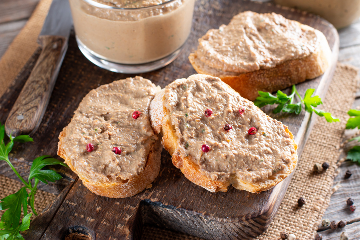 Mehrere Brotscheiben mit Hähnchenleber-Pastete auf einem dunklen Holzbrett, dazu Pfefferkörner und frische Petersilie.