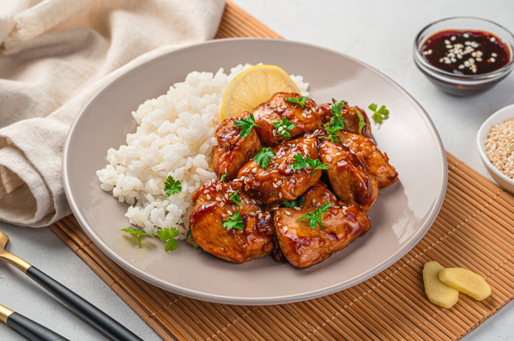 Teriyaki-Hühnchen mit Reis auf einem Teller, dazu glasierte Hähnchenstücke, Sesam, frische Kräuter und eine Zitronenscheibe.