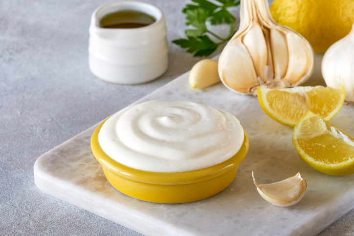 Cremige Aioli in einer gelben Schale auf einer hellen Platte, umgeben von Knoblauch, Zitronenspalten und etwas Öl.