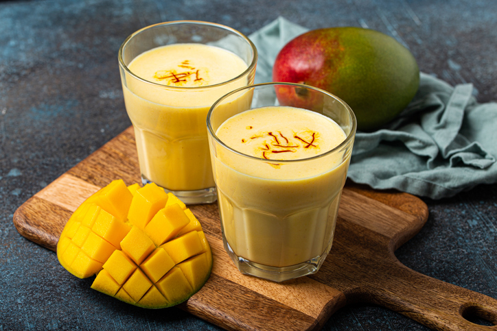 Zwei Gläser Mango Lassi mit Safranfäden auf einem Holzbrett, daneben eine frische Mango und geschnittenes Mangofruchtfleisch.