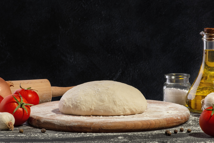 Frischer Pizzateig auf einem bemehlten Holzbrett mit Tomaten, Knoblauch, Olivenöl, Salz und Pfeffer.
