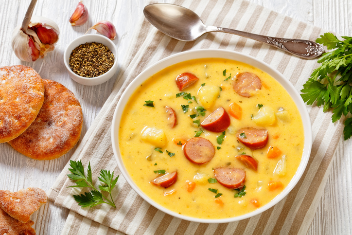 Kartoffelsuppe mit Kartoffelstücken, Wurstscheiben, Möhren und frischen Kräutern in einer weißen Schale auf hellem Tisch.