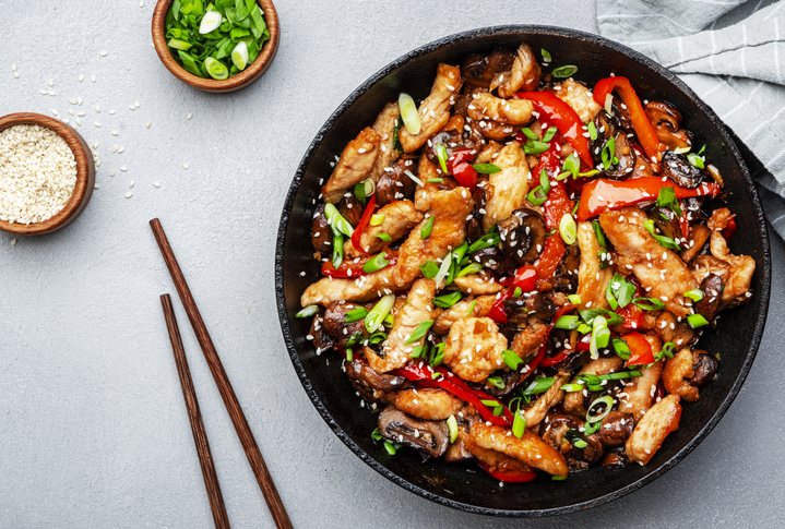 Teriyaki-Hühnchen in einer Pfanne mit Paprika, Pilzen, Frühlingszwiebeln und Sesam auf hellem Untergrund.