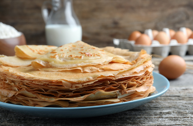 Stapel dünner Crêpes auf einem blauen Teller mit Eiern, Milch und Mehl auf einem rustikalen Holztisch.