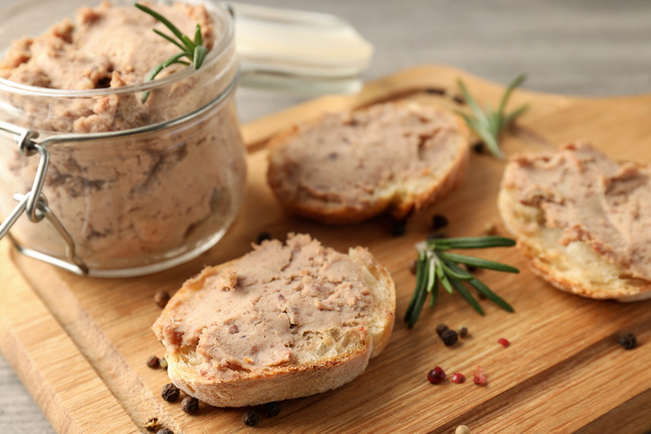 Glas mit Hähnchenleber-Pastete und mehrere bestrichene Brotscheiben auf einem Holzbrett mit Rosmarinzweigen.